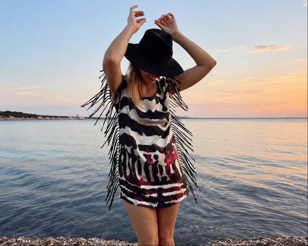 A woman stands on a pebble shore at sunset facing the sea, wearing a black wide-brim hat and a sleeveless fringed dress with a black, white, and red abstract pattern.