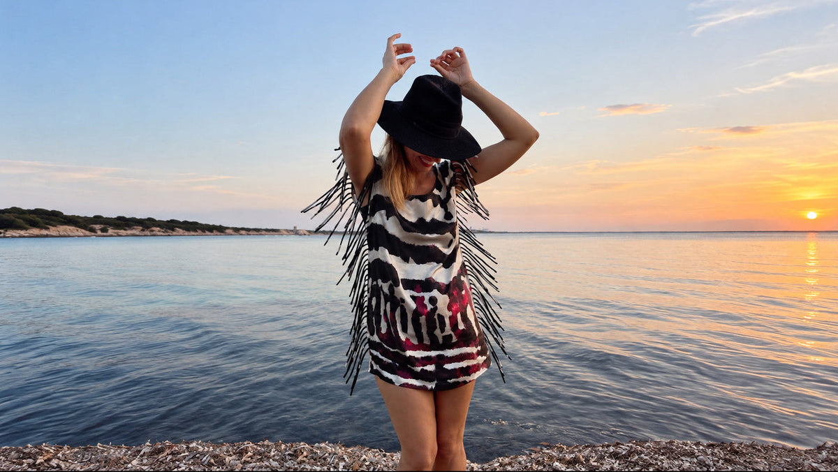 A woman stands on a pebble beach at sunset wearing a black wide-brim hat, a black, white, and red patterned dress with long fringe, and holding a phone to take a photo. The background shows calm water and a glowing orange sunset.