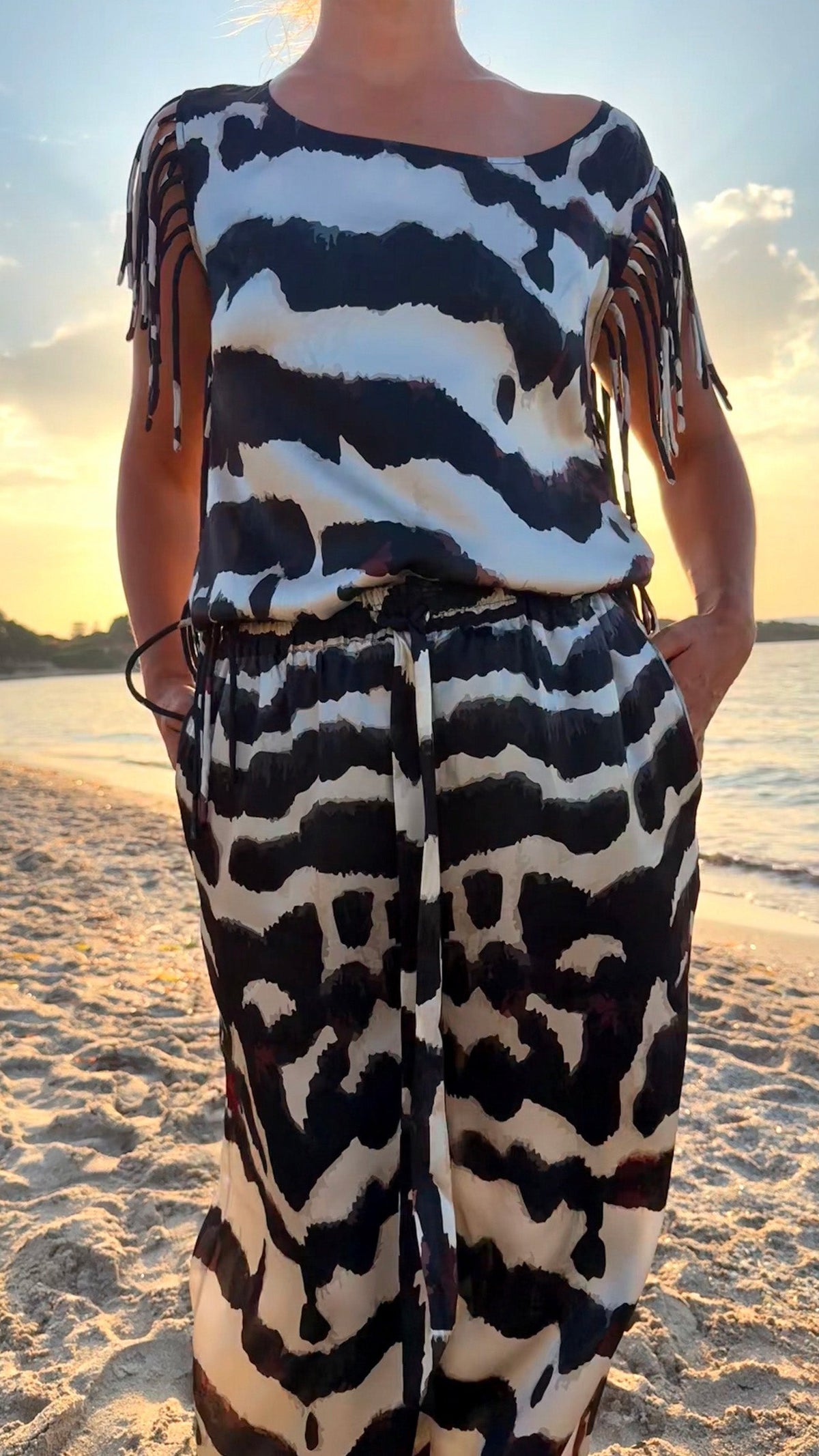 A woman wearing a black and white zebra-print jumpsuit with fringe details on the sleeves, standing on a sandy beach at sunset.