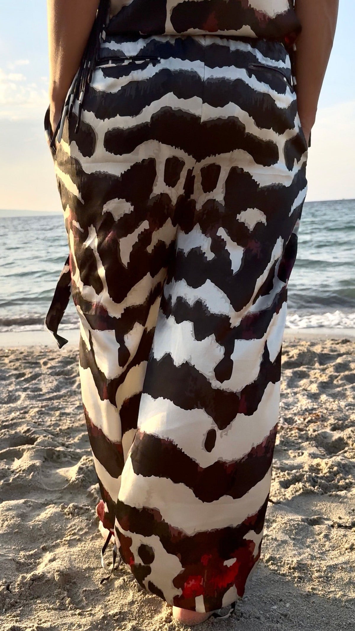 A person standing on a sandy beach wearing a long wrap-style dress or sarong with a bold black, white, and brown abstract animal-inspired pattern. The ocean is visible in the background.