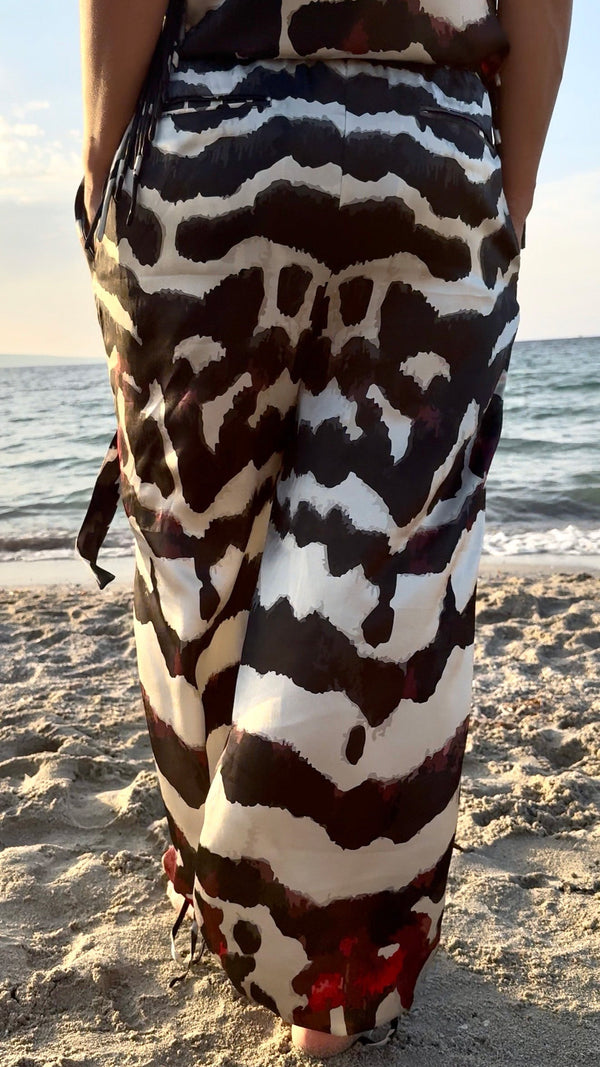 A person standing on a sandy beach wearing a long wrap-style dress or sarong with a bold black, white, and brown abstract animal-inspired pattern. The ocean is visible in the background.